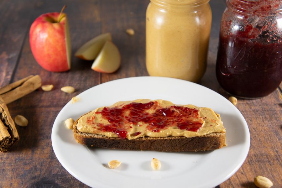 a plate with a slice of bread topped with peanut butter and red jelly in front of an apple and two jars